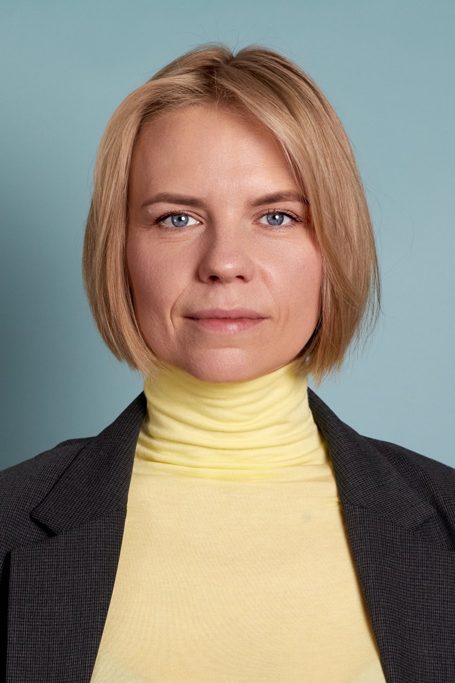 Business portrait of a woman with a blond bob in a yellow turtleneck and dark blazer against a blue backdrop